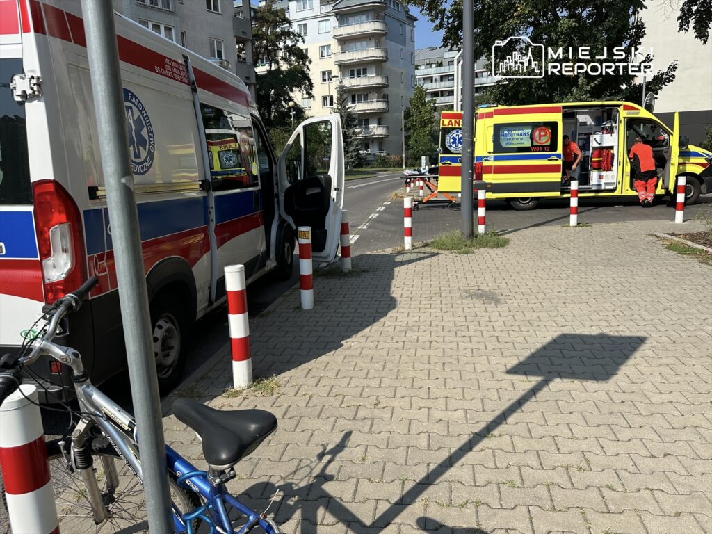 Ambulans z otwartymi drzwiami zaparkowany obok drugiego ambulansu, na chodniku w miejskim otoczeniu.