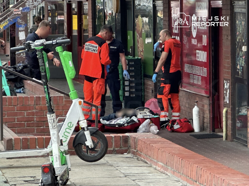 Ratownicy medyczni udzielają pomocy osobie leżącej na chodniku przed sklepem, obok skuterów elektrycznych.