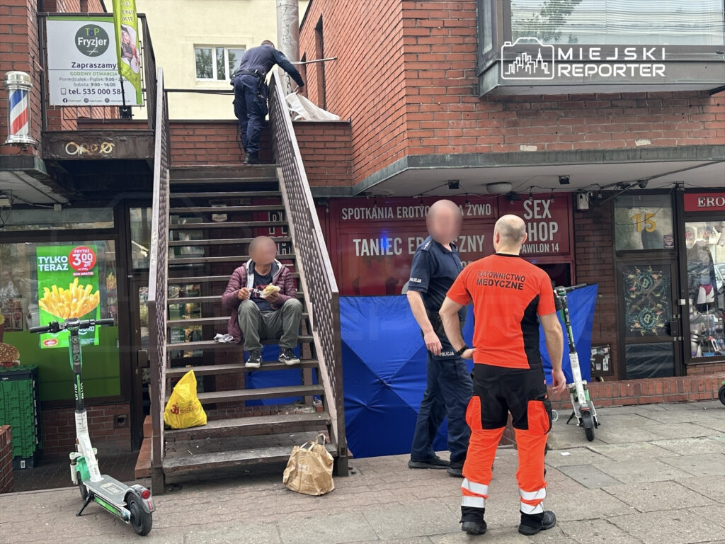 Mężczyzna siedzący na schodach, jedzący jedzenie, podczas gdy policjant i ratownik medyczny rozmawiają obok.