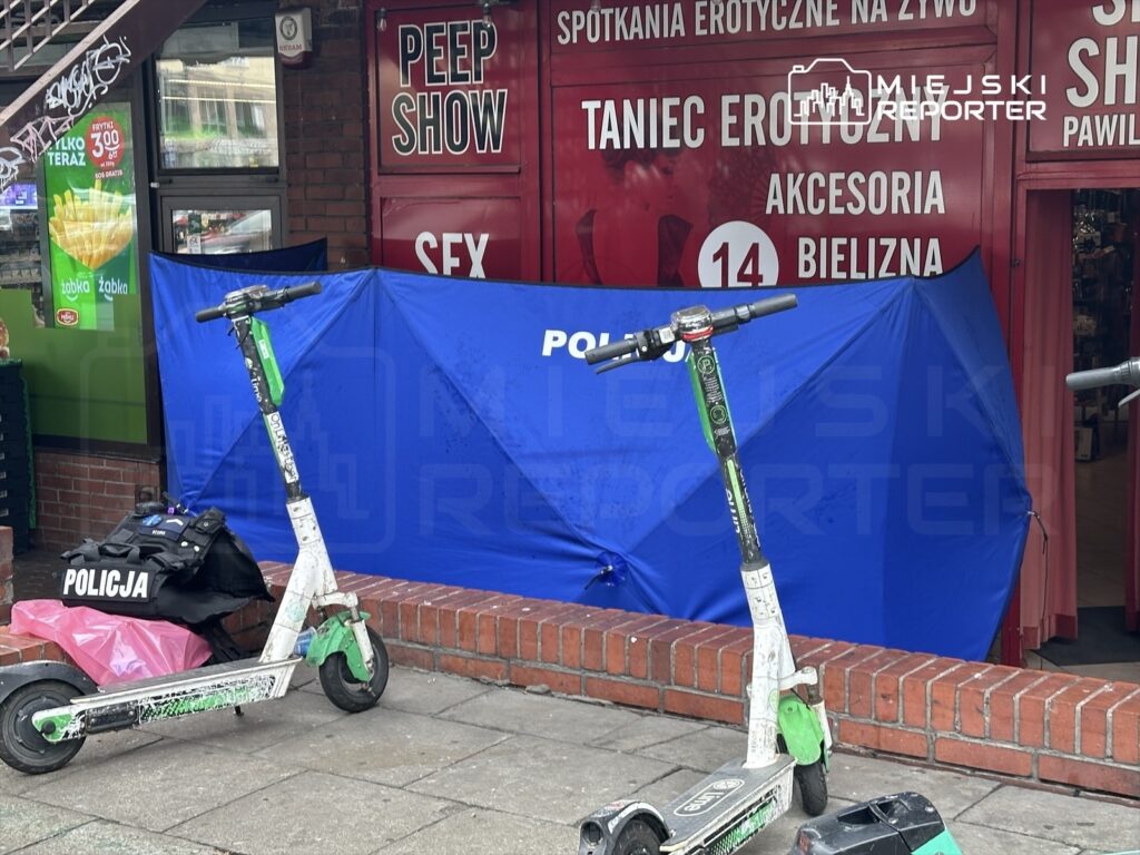 Policjanci zabezpieczają miejsce zdarzenia za niebieską plandeką, obok stoją elektryczne hulajnogi na chodniku.