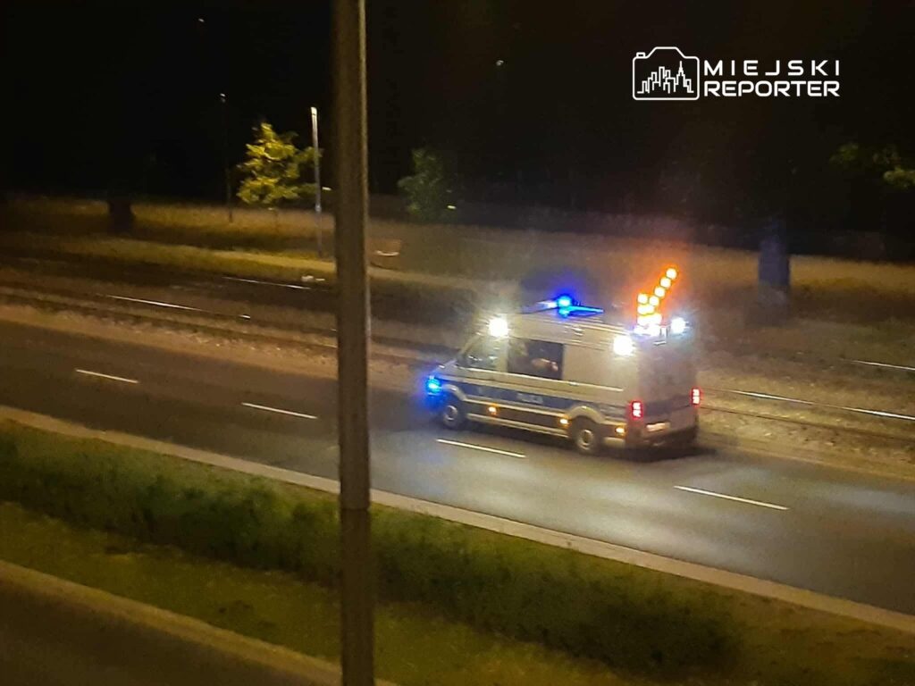 Radiowóz policyjny z włączonymi sygnałami świetlnymi porusza się po nocnej drodze obok torów kolejowych.