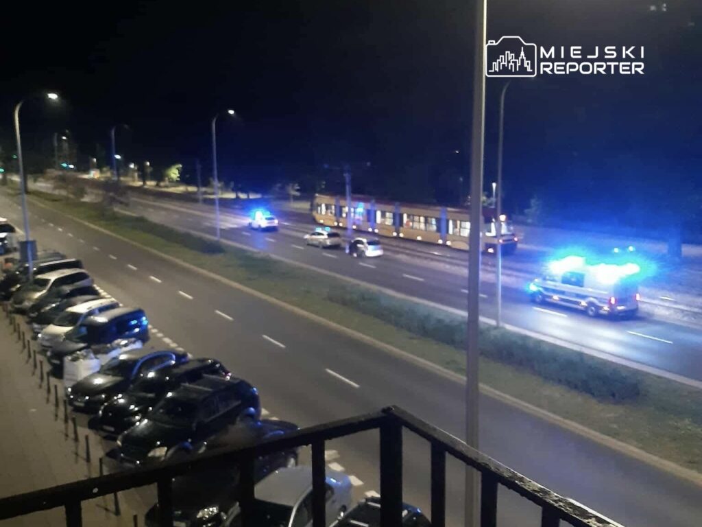 Na ulicy w nocy porusza się tramwaj obok radiowozu i karetki, z włączonymi sygnalizacjami świetlnymi.