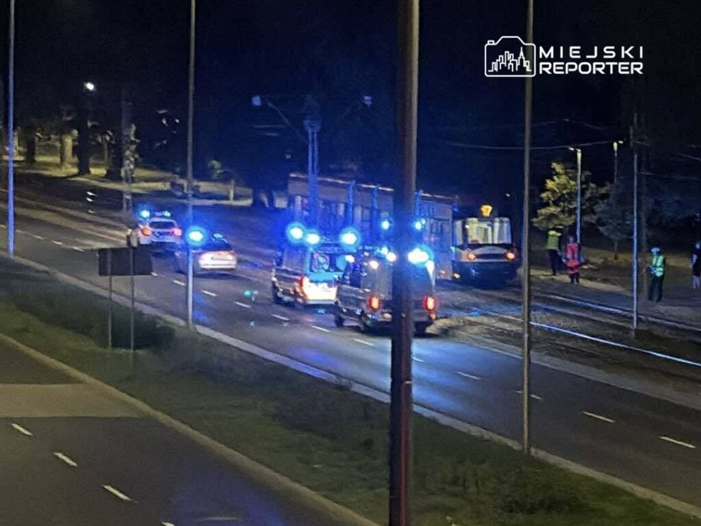 Na jezdni nocą zatrzymały się pojazdy ratunkowe z włączonymi sygnałami świetlnymi, obok stoi tramwaj.