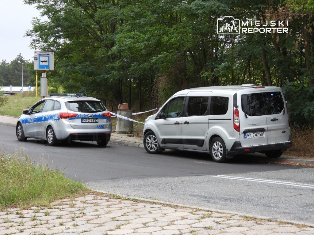 Policyjny samochód osobowy i van zaparkowane obok przystanku autobusowego, z taśmą zabezpieczającą w tle.