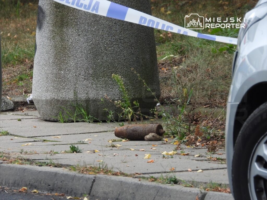 Na chodniku leży metalowy przedmiot przypominający pocisk, obok słupa ogrodzonego taśmą policyjną.