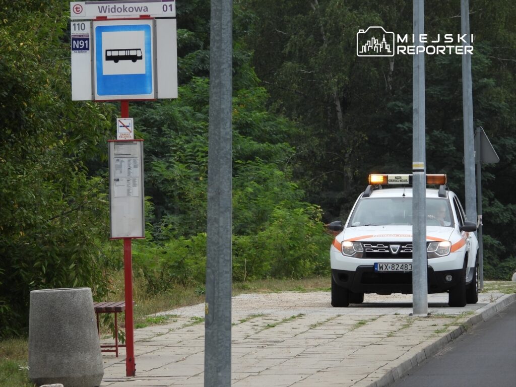 Pojazd służbowy zaparkowany przy przystanku autobusowym w otoczeniu zieleni.