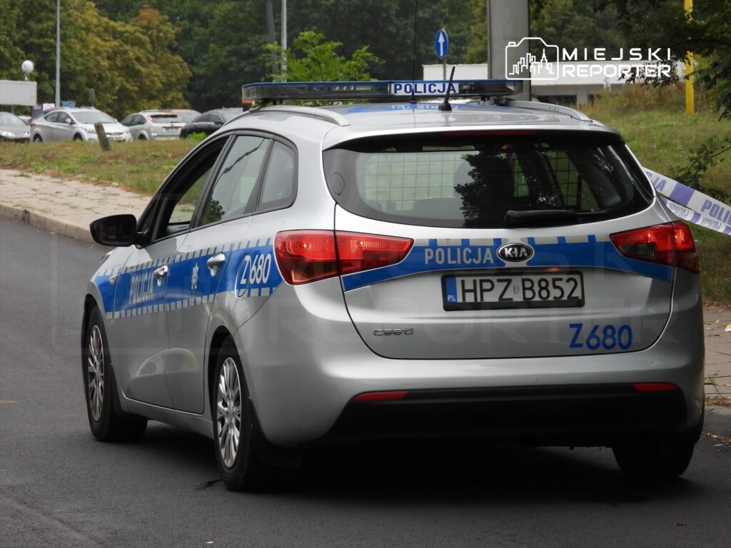 Policyjny samochód marki Kia z niebieskimi oznaczeniami, zaparkowany na ulicy w otoczeniu zieleni.