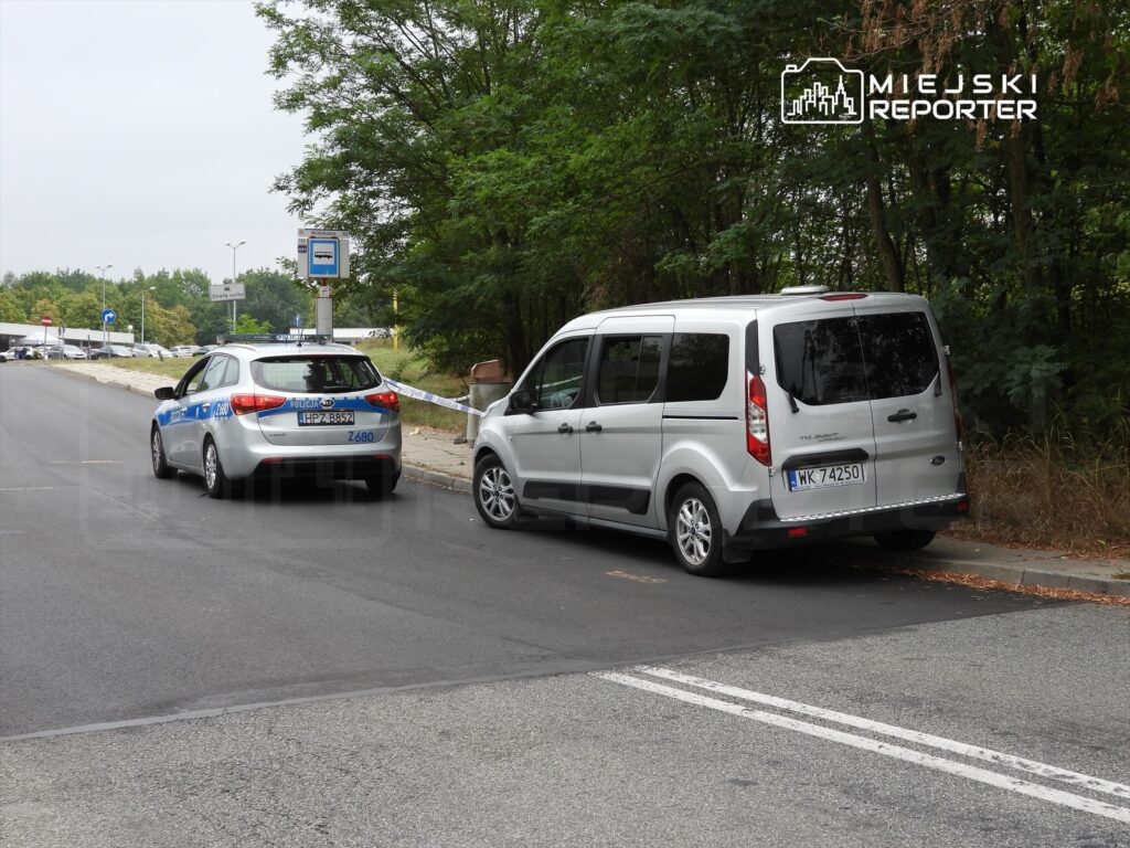Policyjny radiowóz zatrzymany obok szarego vana na ulicy, w tle zieleń i przystanek autobusowy.