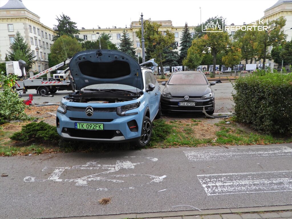 Niebieski samochód osobowy z otwartą maską stoi na chodniku obok czarnego pojazdu, w tle widać prace porządkowe.