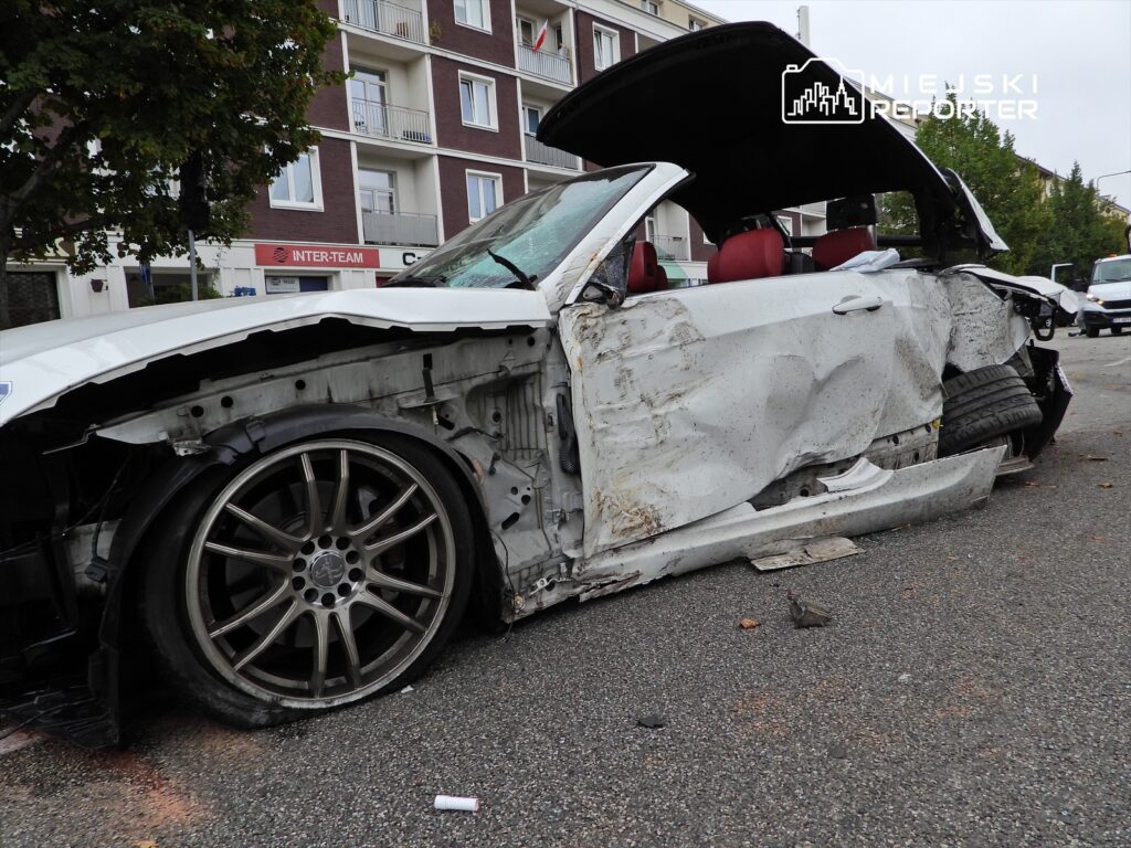 Uszkodzone białe auto sportowe z otwartym dachem stoi na ulicy, z widocznymi uszkodzeniami karoserii i jednym odkręconym kołem.