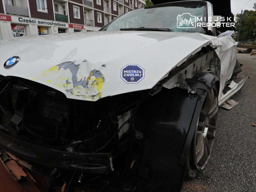 Uszkodzony biały samochód BMW z wgniecioną maską i naklejką „MISTRZU ZWOLNIJ” na parkingu.