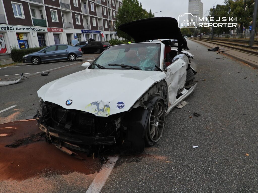 Uszkodzony biały samochód marki BMW stoi na jezdni, obok widać inne pojazdy oraz ślady wypadku.