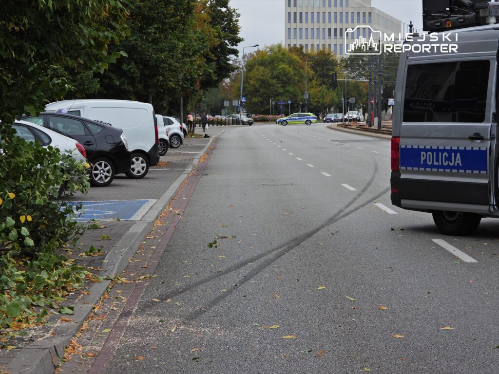 Na ulicy zaparkowane samochody, w tle radiowóz policji oraz zniszczona nawierzchnia z liśćmi.