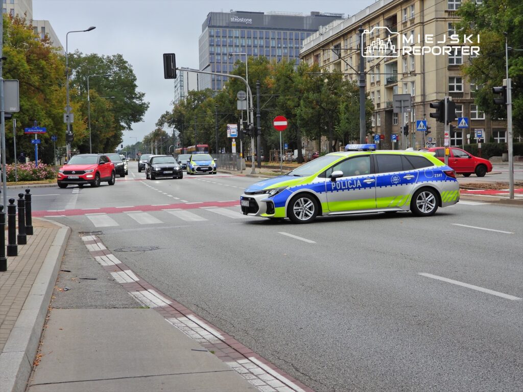 Radiowóz policyjny zatrzymany na skrzyżowaniu, obok poruszające się samochody osobowe w miejskim otoczeniu.