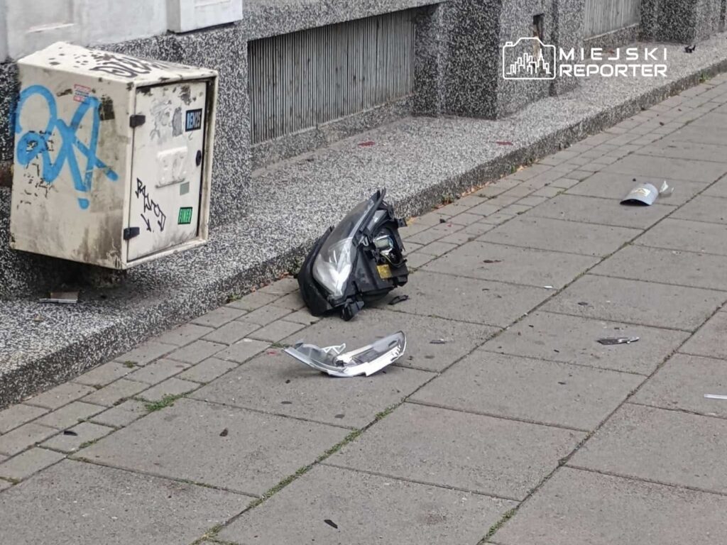 Na chodniku leży uszkodzony element samochodowy obok skrzynki z graffiti. W tle widać szary budynek.