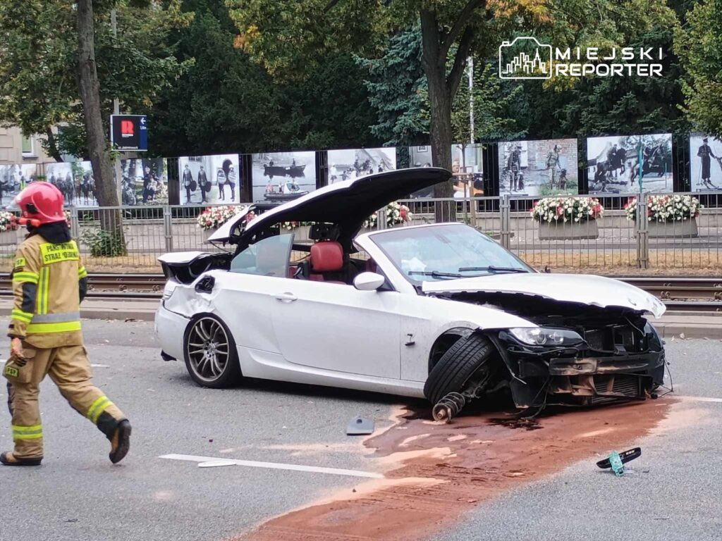 Uszkodzony biały samochód sportowy stoi na jezdni, obok przechodzi strażak w hełmie.