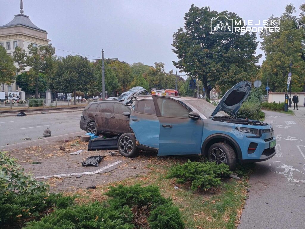Zniszczony samochód osobowy z otwartą maską stoi na chodniku, obok uszkodzonego pojazdu i porozrzucanych elementów.