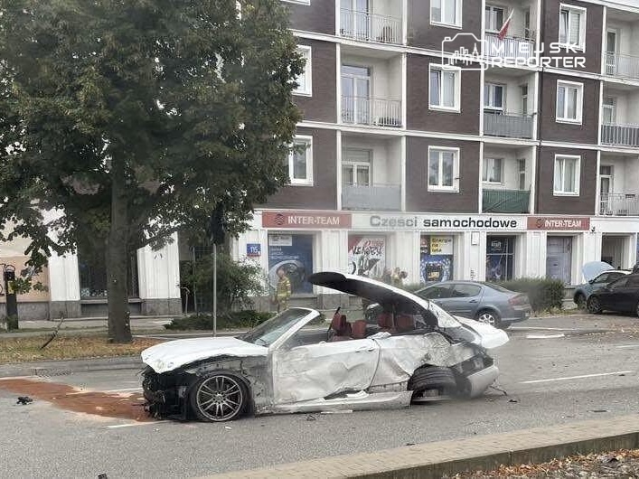 Zniszczony samochód osobowy z otwartym dachem leży na drodze obok budynku mieszkalnego.