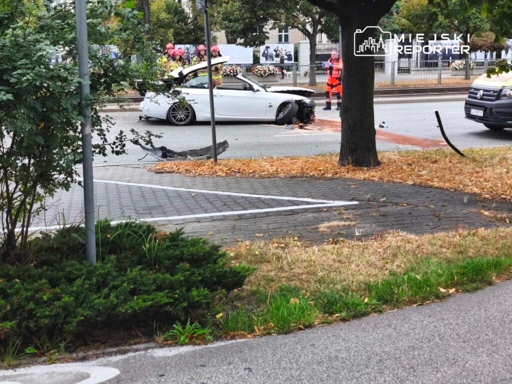 Biały samochód osobowy z uszkodzoną przednią częścią stoi na jezdni, obok grupy strażaków w kaskach.