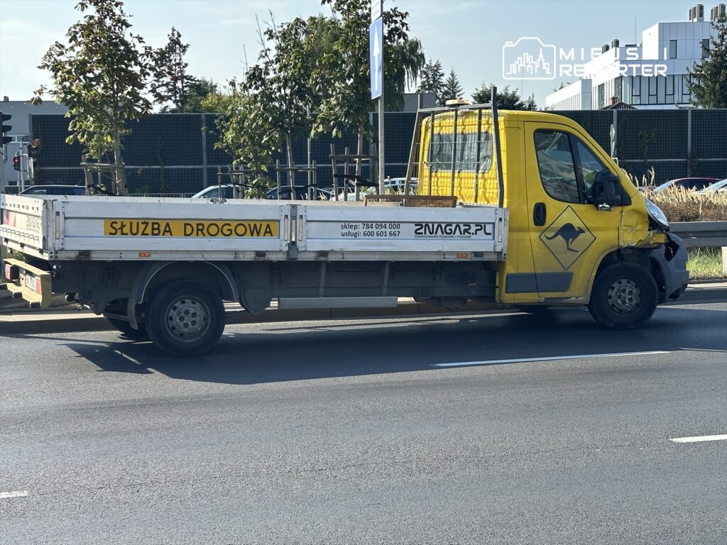 Żółty samochód dostawczy z napisem "SŁUŻBA DROGOWA" zatrzymany na ulicy, obok drzew i nowoczesnych budynków.