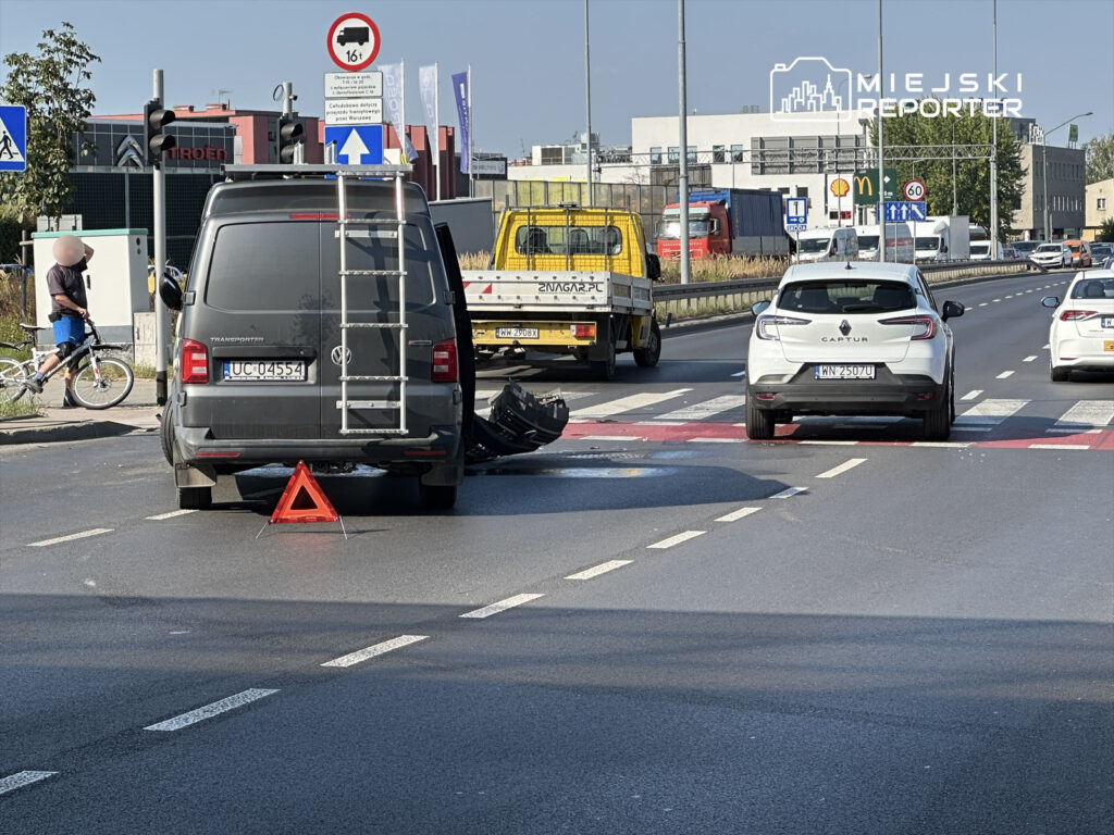Na drodze stoi czarny van z włączonym trójkątem ostrzegawczym, obok zderzona biała osobówka. W tle widoczne inne pojazdy.