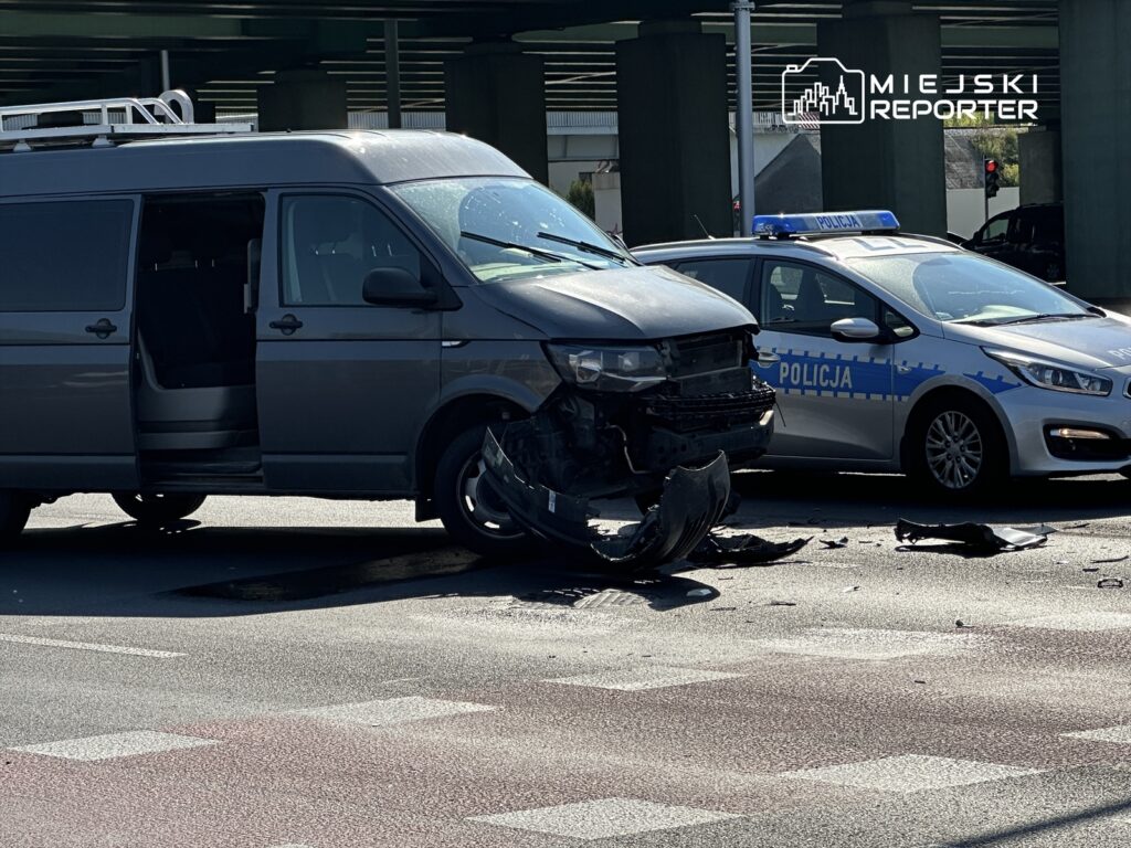 Uszkodzony szary van z otwartymi drzwiami stoi na drodze, obok niego policyjny radiowóz z napisem "POLICJA".