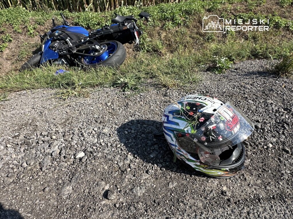 Motocykl leżący na poboczu drogi obok kasku, w otoczeniu zieleni i żwirowej nawierzchni.