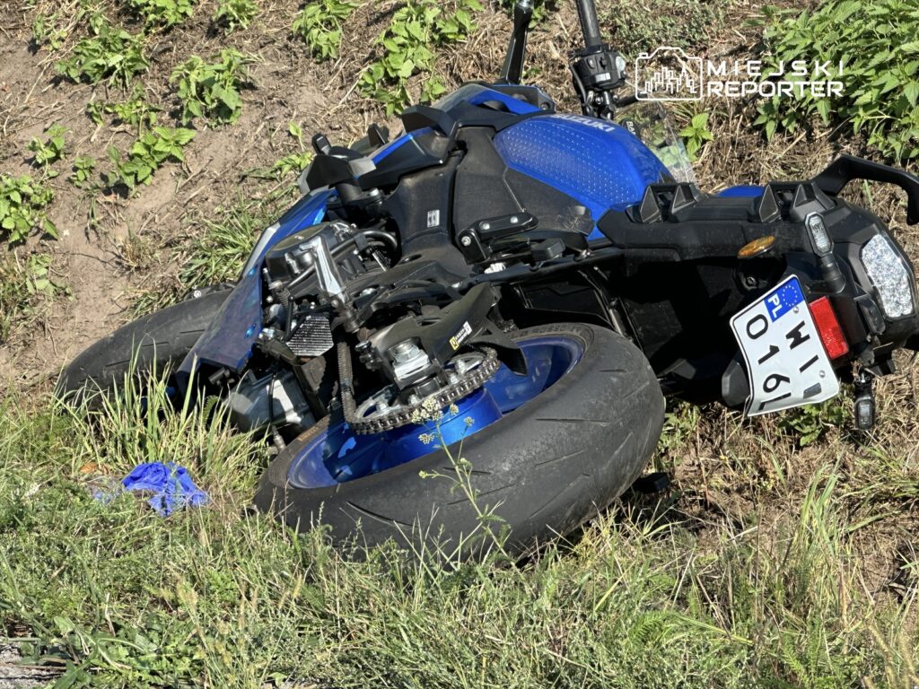 Niebieski motocykl leży na boku w trawie obok drogi, z widocznym uszkodzeniem.