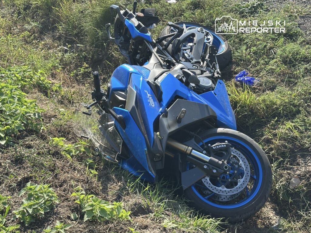 Niebieski motocykl leży na boku wśród trawy i chwastów, z uszkodzoną konstrukcją.