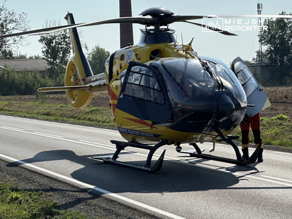 Śmigłowiec ratunkowy w żółto-czerwonych barwach lądował na poboczu drogi, obok stoi ratownik w czerwonym stroju.