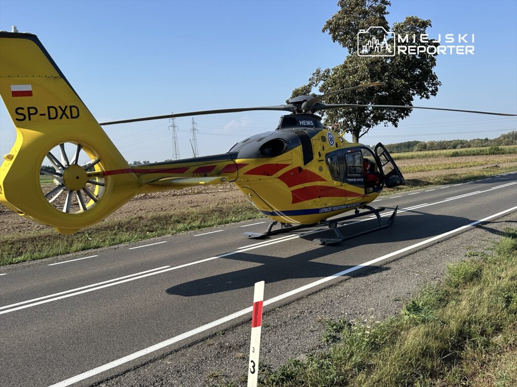 Żółty śmigłowiec ratunkowy lądował na drodze, obok pola, w słoneczny dzień.