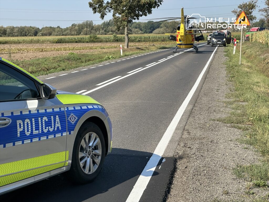 Policyjny radiowóz z niebieskimi i żółtymi oznaczeniami stoi na drodze, obok lądowiska dla helikoptera.