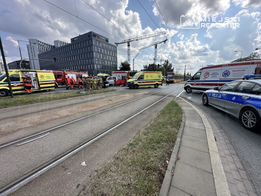 Zespół ratunkowy oraz strażacy pracują na miejscu wypadku w pobliżu nowoczesnego biurowca, obok licznych pojazdów służb.