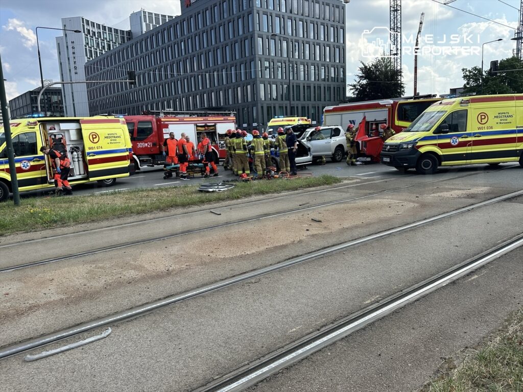 Zespół ratunkowy w pomarańczowych strojach pracuje przy wypadku drogowym, obok uszkodzonych pojazdów i wozów strażackich.