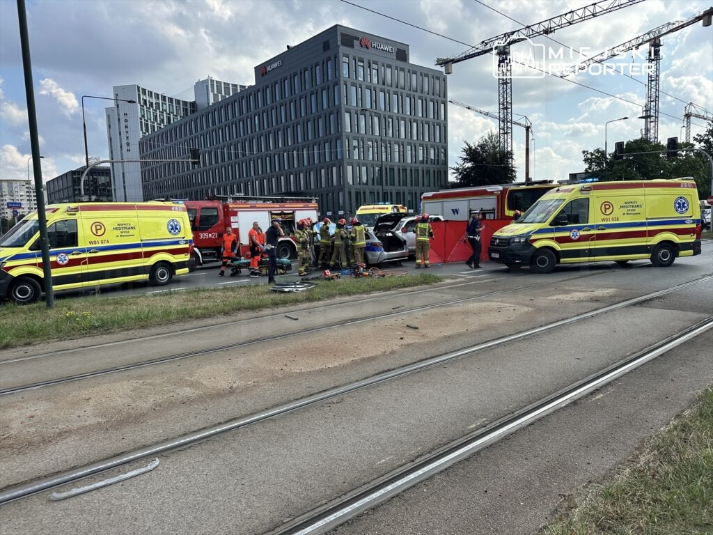 Zespół ratunkowy pracuje nad wypadkiem drogowym, obok stoją karetki i wóz strażacki, w tle nowoczesne budynki.