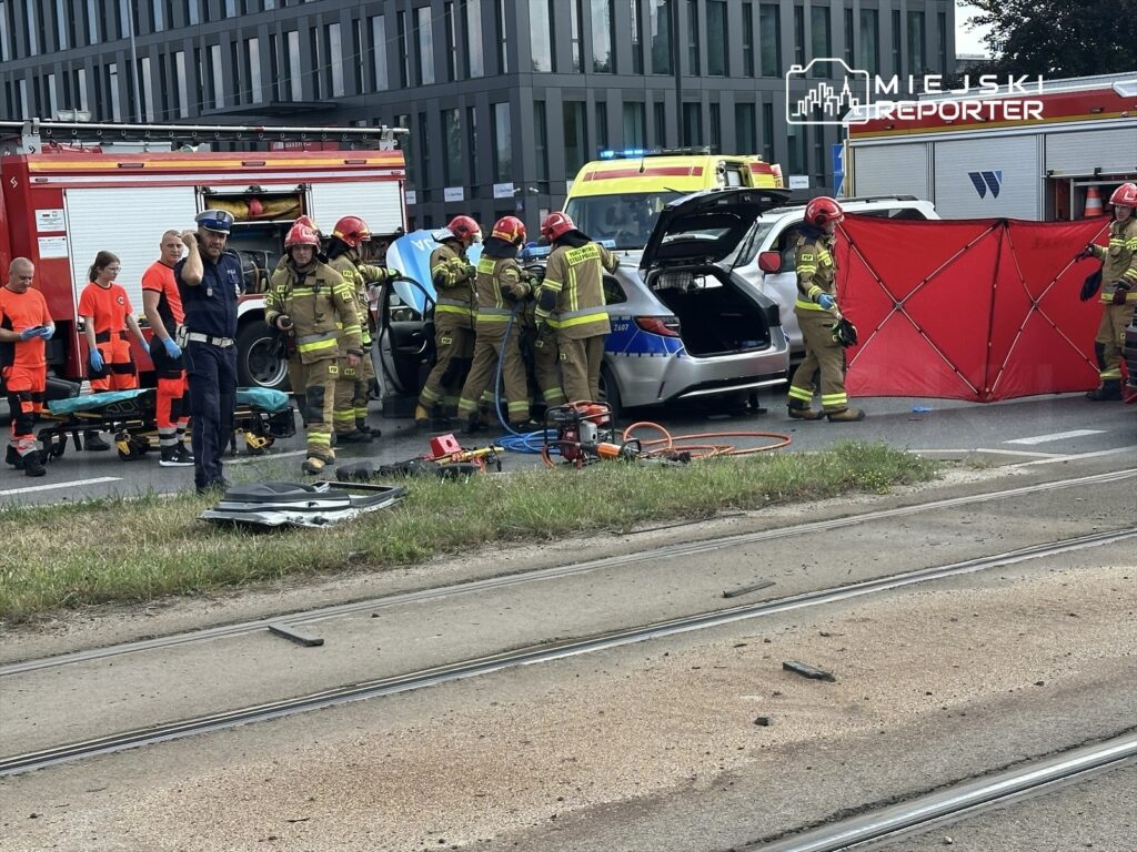 Grupa strażaków w hełmach pracuje przy samochodzie osobowym, obok znajduje się karetka i wóz strażacki.