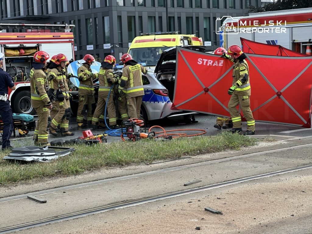 Grupa strażaków w mundurach ratunkowych prowadzi akcję ratunkową przy rozbitej karecie w miejskim otoczeniu.