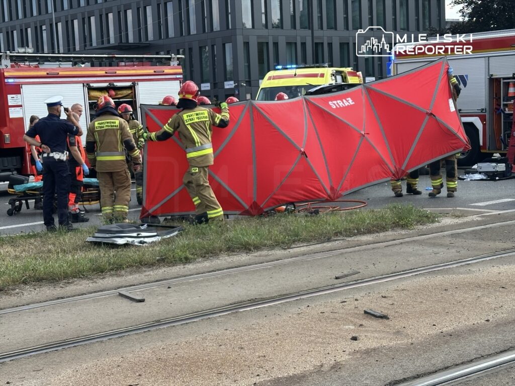 Grupa strażaków ustawia czerwoną osłonę w pobliżu wypadku drogowego, obok karetki i wozu strażackiego.