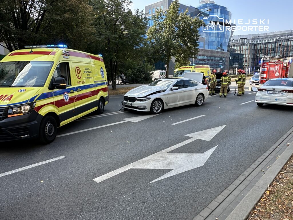 Ambulans i samochód osobowy stoją na ulicy, obok grupy strażaków zajmujących się interwencją.