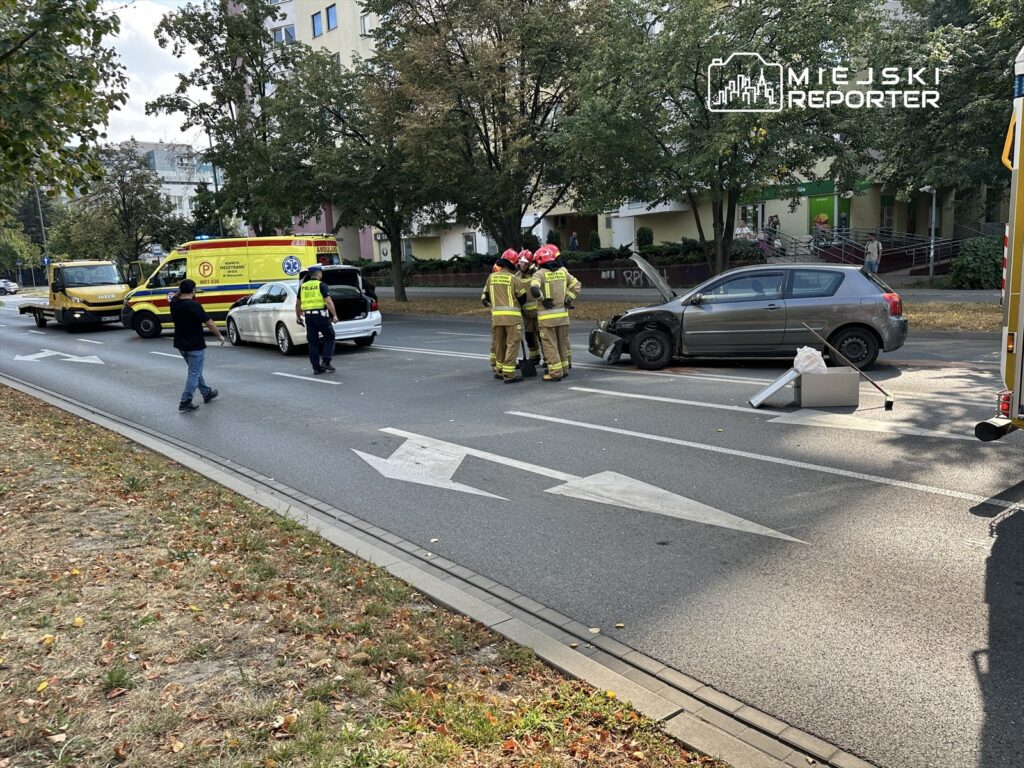 Grupa strażaków w hełmach omawia sytuację przy uszkodzonym samochodzie na ulicy, obok parkującego ambulansu.