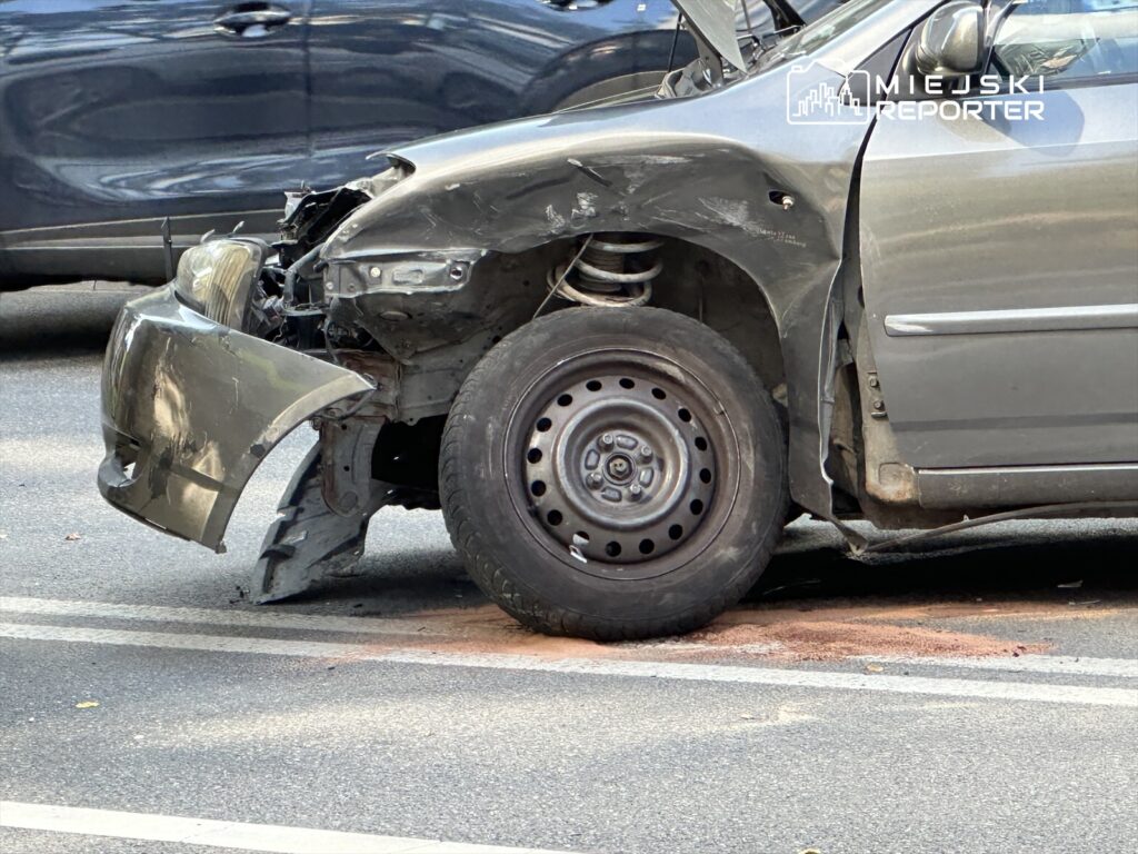 Uszkodzony samochód z zdemolowanym przodem stoi na drodze, obok widoczne inne pojazdy. Na jezdni plama płynu.