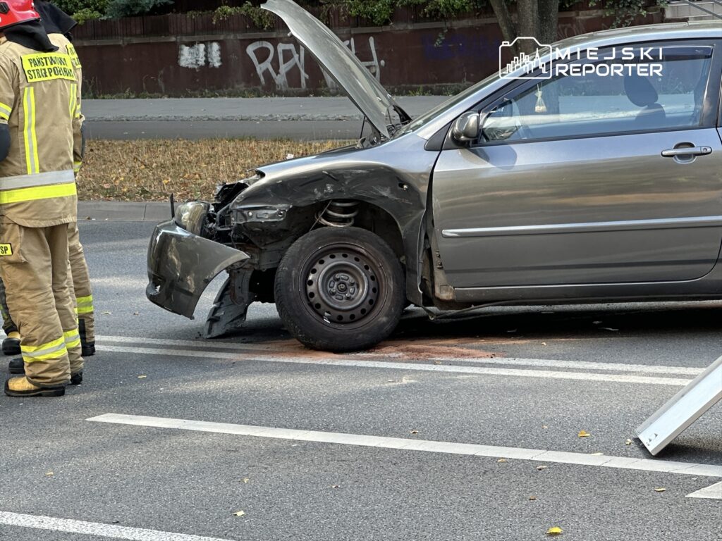 Strażacy w mundurach rozmawiają obok uszkodzonego samochodu z otwartą maską na ulicy.