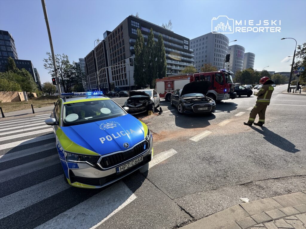 Policjant przy radiowozie zabezpiecza miejsce wypadku drogowego z udziałem dwóch rozbitych samochodów.