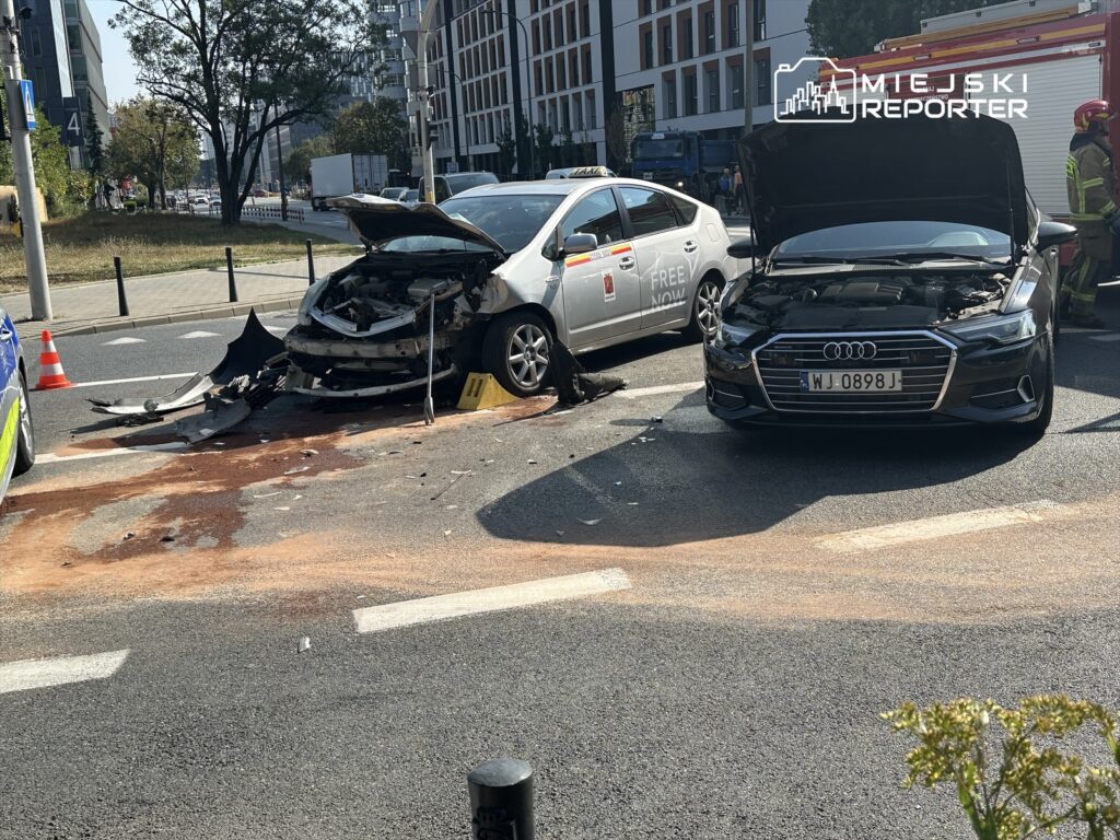 Dwa uszkodzone samochody na ulicy, jeden z otwartą maską, a obok leży fragment karoserii. W tle widać ruch uliczny.