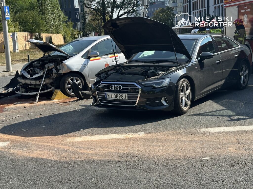Wypadek drogowy z udziałem uszkodzonego srebrnego samochodu z otwartą maską i czarnego Audi na drodze.