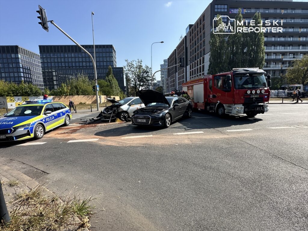 W wyniku wypadku drogowego na skrzyżowaniu stoi uszkodzony samochód osobowy, obok policja i straż pożarna.