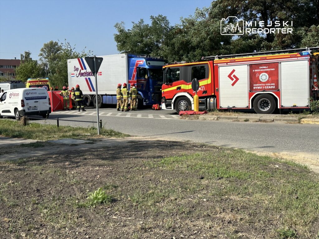 Grupa strażaków w kaskach i mundurach pracuje przy wypadku drogowym z udziałem ciężarówki i ambulansu na miejskiej ulicy.