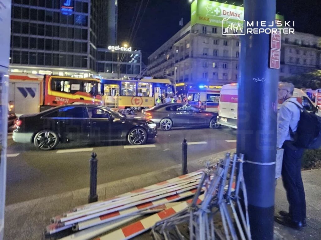 Czarne samochody osobowe stoją w korku obok tramwaju w nocy, w tle widać niebieskie światła służb ratunkowych.