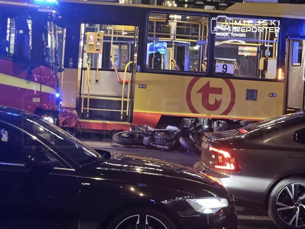 W nocy, w miejskim otoczeniu, autobus z włączonymi sygnalizatorami świetlnymi stoi obok przewróconego motocykla, otoczony przez samochody osobowe.