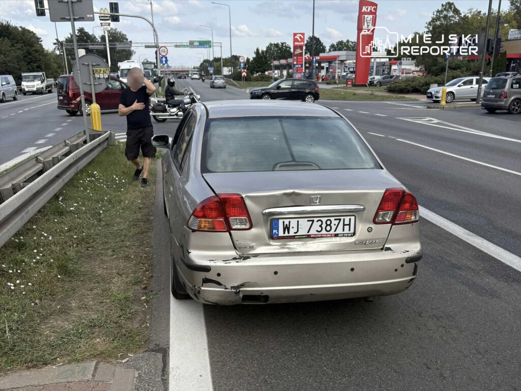 Mężczyzna w czarnej koszulce idzie wzdłuż drogi obok uszkodzonego samochodu na pasie ruchu.